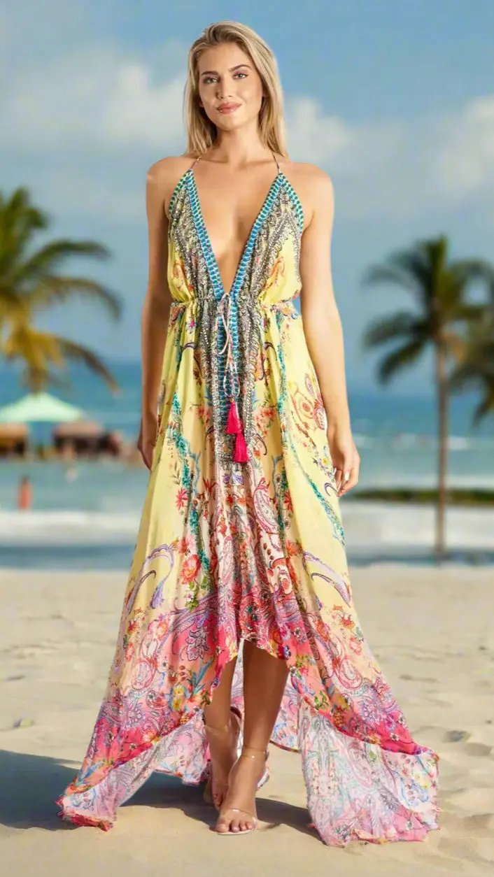 On a sunny beach with palm trees and ocean views, a woman wears the La Moda Clothing Resortwear Maxi Halter Dress for Summer Vacations—a flowing sundress in vibrant yellow, pink, and blue with a deep V-neck, intricate patterns, and tassel accents.