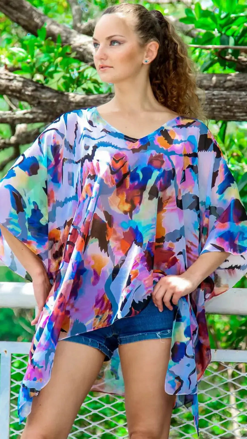 A woman with curly hair confidently poses outdoors in a vibrant, loose-fitting lamodaclothings Sheer Kaftan-Style Cover-Up layered over blue denim shorts, hands on hips against greenery and a white fence.