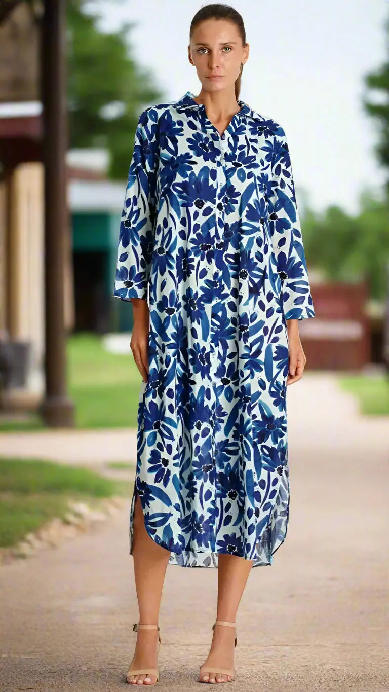 A woman wears the La Moda Clothing Shirt Dress with Pockets—a long, button-up style featuring a blue and white floral pattern, three-quarter sleeves, and a collar—outdoors with her hair tied back and nude heels. Green trees and buildings blur in the background.