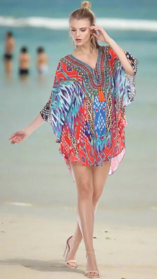 A woman with light skin and blonde hair tied back walks on the beach in Lamodaclothing's Short Balloon Kaftan Luxury Beachwear. She pairs it with nude heels as the ocean and blurred beachgoers appear in the background.