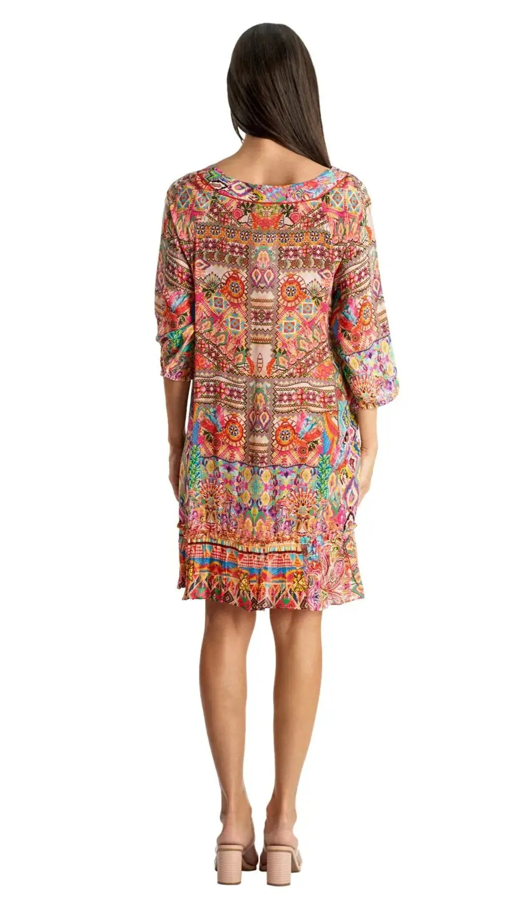 A woman with straight dark hair stands with her back to the camera wearing the La Moda Clothing Short Dress—knee-length, loose, and vibrant with multicolored geometric and floral patterns—paired with light heeled sandals.