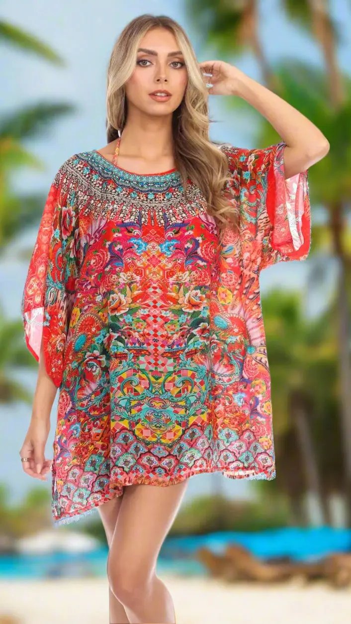 A woman stands on a beach amid palm trees, wearing the Soul of Summer Kaftan Top by La Moda Clothings—a colorful, loose knee-length kaftan with bold red, blue, and green patterns. She has long blonde wavy hair and a neutral expression.