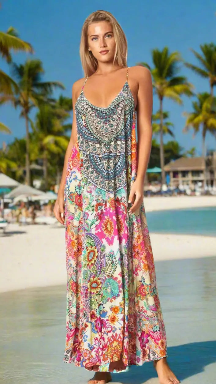A blonde woman stands barefoot on a beach in a Lamodaclothing Spaghetti Strap Maxi Dress with Front Pockets. Palm trees, blue sky, ocean, and distant lounge chairs are visible in the background.