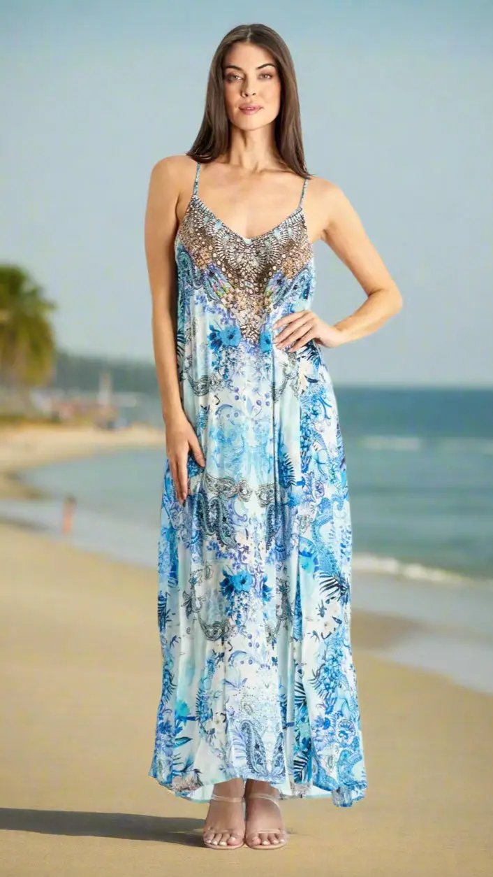 On a sandy beach with ocean and palm trees, a woman with long brown hair and light skin poses in Lamodaclothing's Spaghetti Strap Maxi Resort Dress with Front Pockets—a flowing, blue and white patterned sleeveless dress.