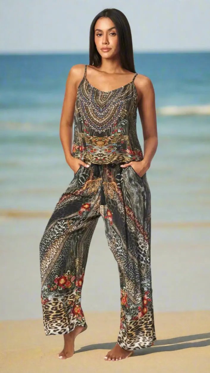 A woman in the La Moda Clothing Summer Camisole Top with Pants Outfit stands barefoot on the beach, hands in her pockets, wearing wide-leg, mixed animal print pants and a loose top as she gazes confidently at the camera.
