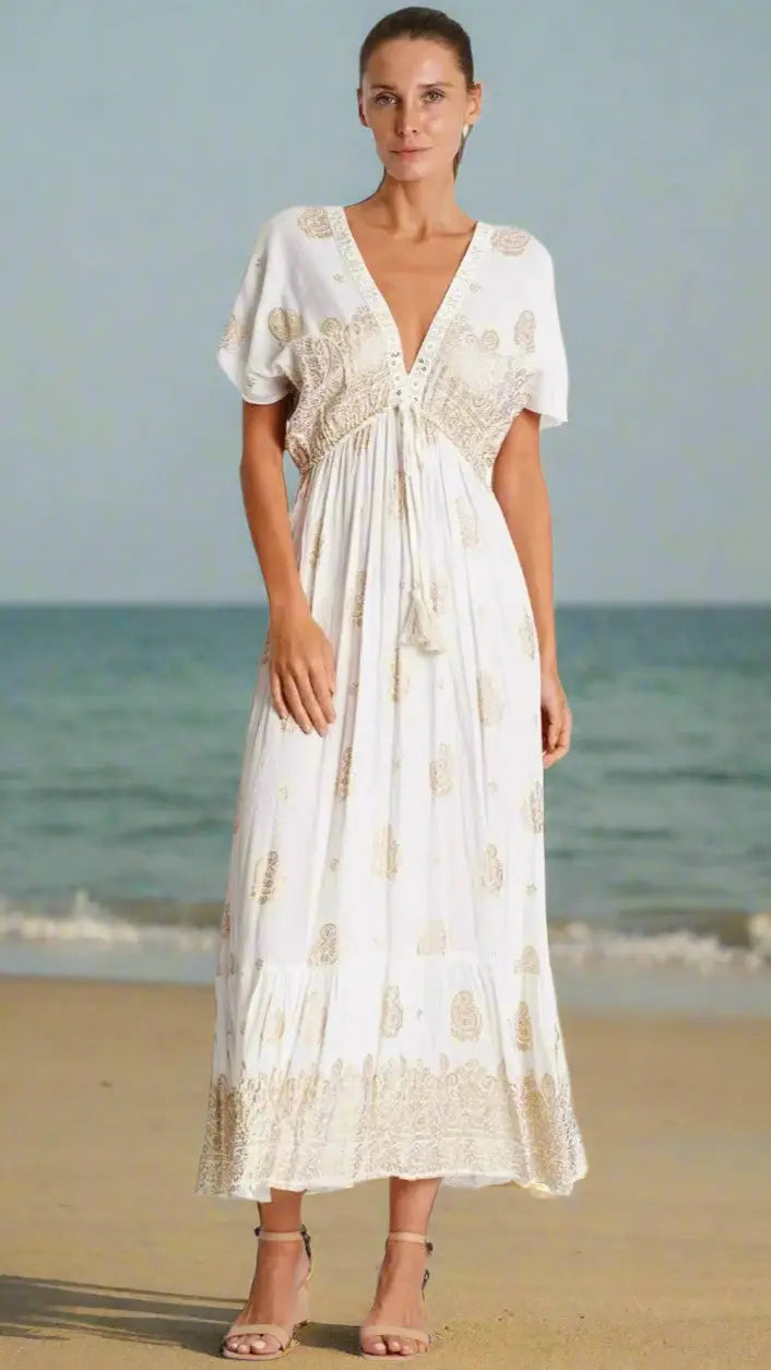 A woman stands barefoot on the beach in a La Moda Clothing Summer Dress—white, ankle-length, with short sleeves, a deep V-neck, and tan paisley patterns. Her hair is pulled back and she looks calm with the sea behind her.