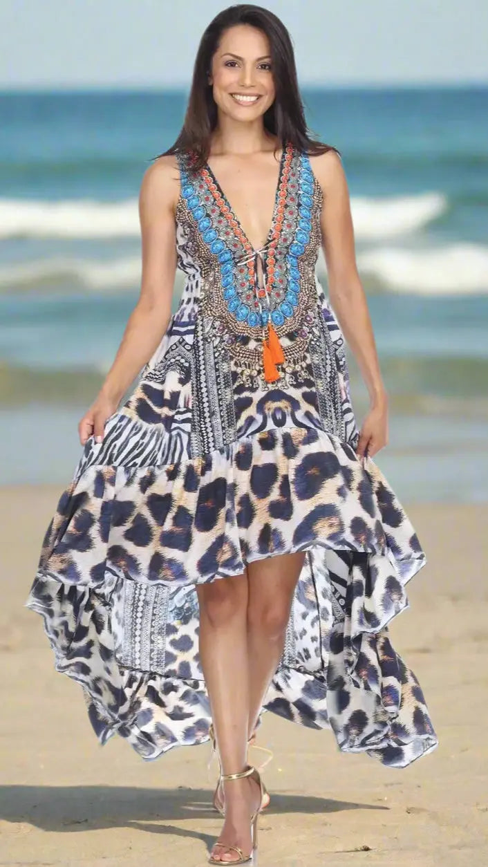 A woman with long dark hair walks on the beach, smiling in a Lamodaclothing Summer Evening Casual Dress for Resort Cruise Vacation—sleeveless, high-low, featuring animal prints, blue embroidery, orange tassels—paired with strappy heels. Waves and sand behind her.