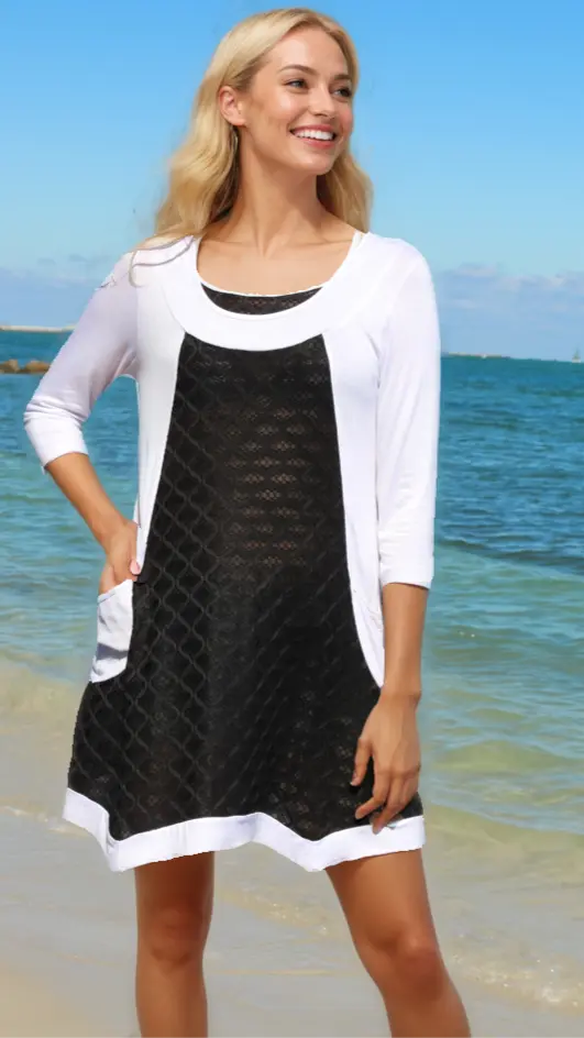 A smiling woman with long blonde hair stands on a beach in the Lamodaclothing Summer Fashion Resort Wear Tunic dress, featuring three-quarter sleeves and a black geometric lace front panel. She looks to the side with one hand in her pocket under a clear blue sky.