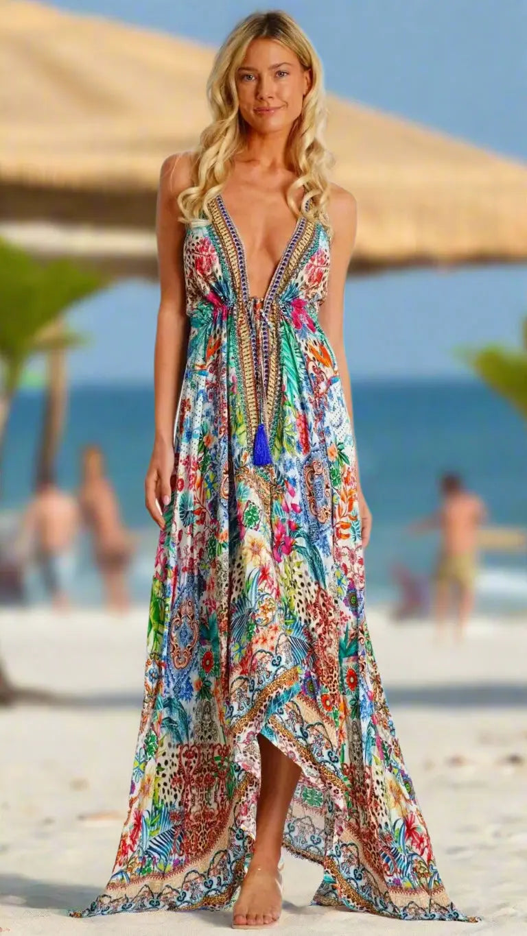 A smiling woman with long blonde hair wears the La Moda Clothing Summer Halter Dress, a colorful, flowy maxi with tassels and a deep V-neck. She stands barefoot on a sandy beach, with palm trees and the sea blurred in the background.
