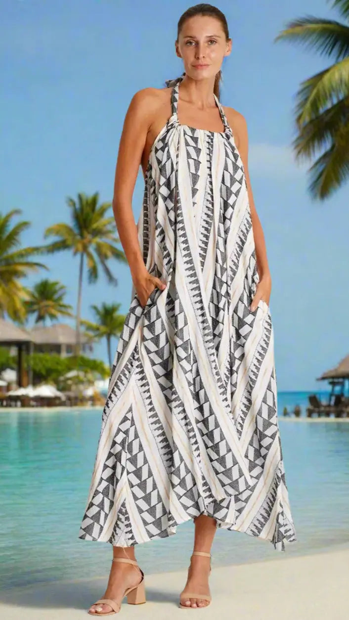 A woman stands by a tropical beach in the La Moda Clothing Summer Halter Neck Dress, a sleeveless black and white geometric maxi with side pockets. She pairs it with nude heeled sandals, palm trees and thatched roofs in the background.