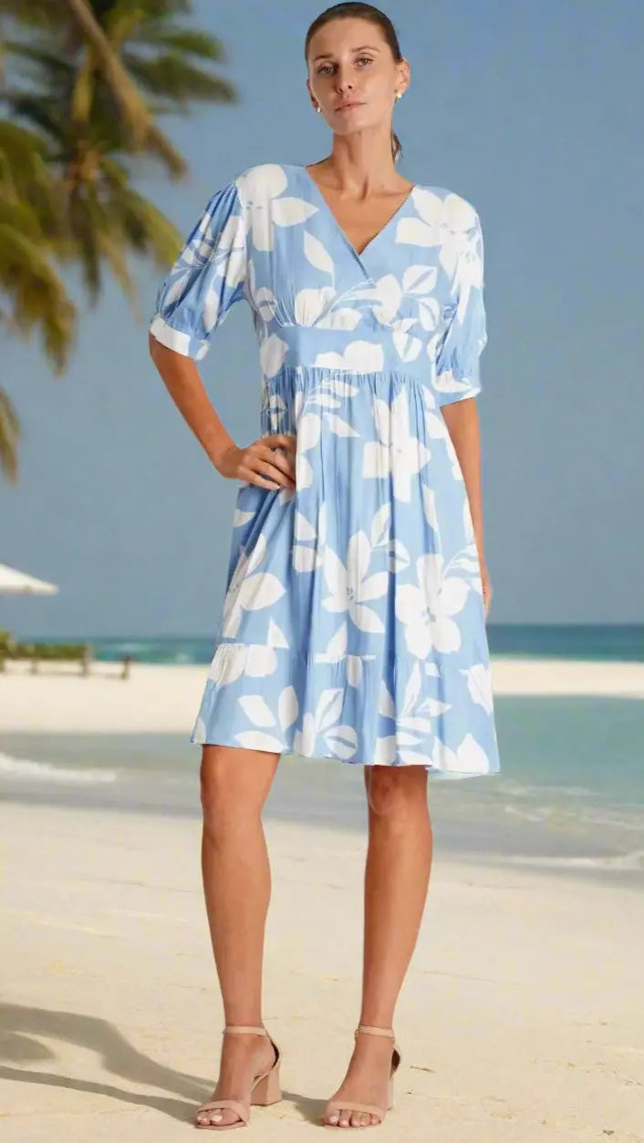 A woman stands barefoot on a sandy beach with palm trees, wearing the La Moda Clothing Summer Mini Dress featuring light blue fabric, large white floral patterns, short puff sleeves, and a V-neck. The background shows a clear sky and calm ocean.