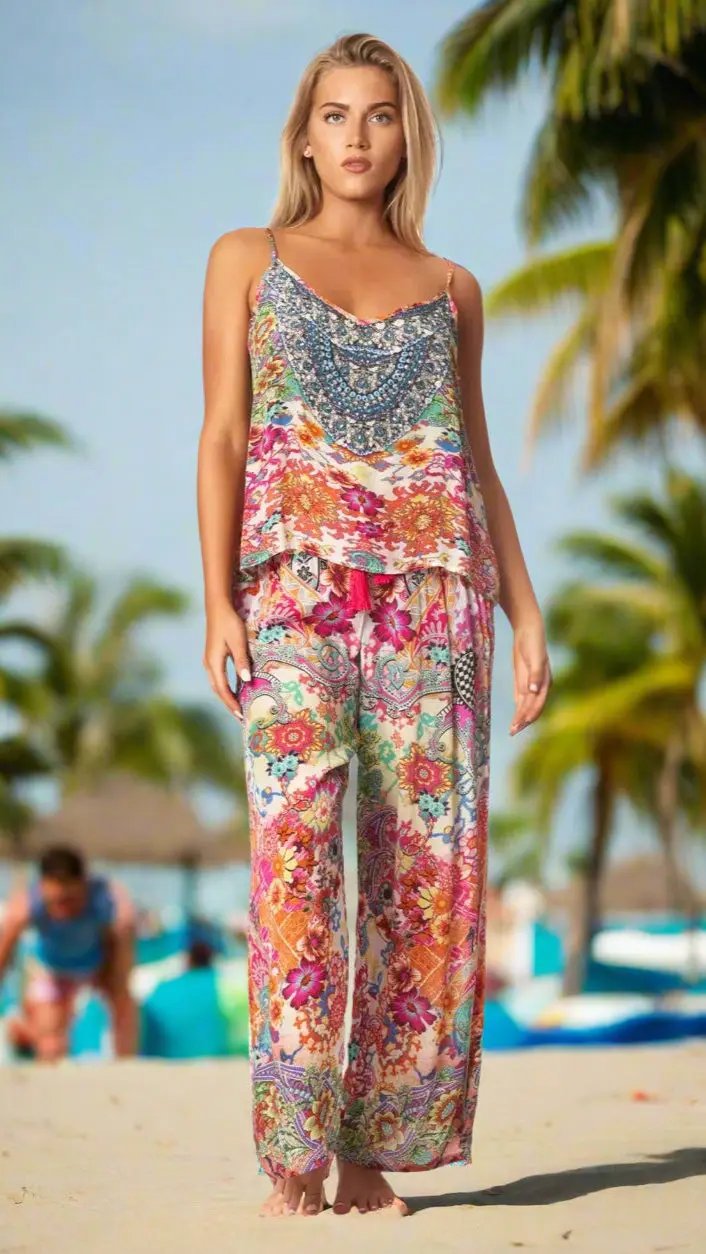 A woman stands barefoot on a sandy beach, wearing the La Moda Clothing Summer Vacation Lifestyle Pant Set. Palm trees and blurred beachgoers are visible in the background under a clear sky.