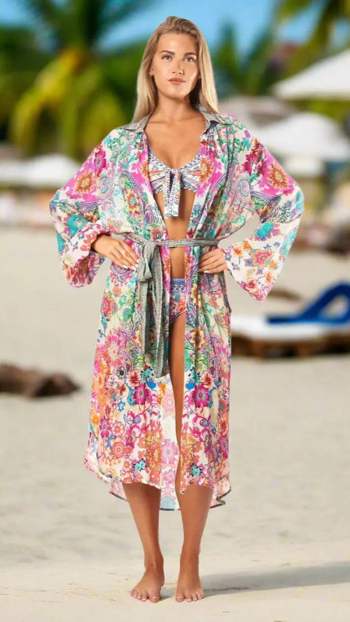 A woman stands barefoot on a sandy beach, confidently wearing The Effortless Shirtdress Cover-Up by La Moda Clothings over her bikini. Palm trees and lounge chairs blur in the background as sunlight brightens her face and hair.
