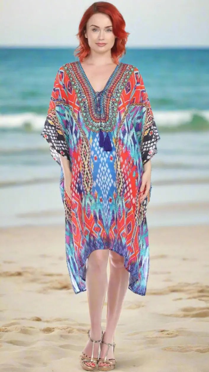 A red-haired woman stands barefoot on the sand by the ocean, smiling in a colorful Trendy V-Neckline Luxury Kaftan Dress with blue and red geometric patterns from Lamodaclothing.