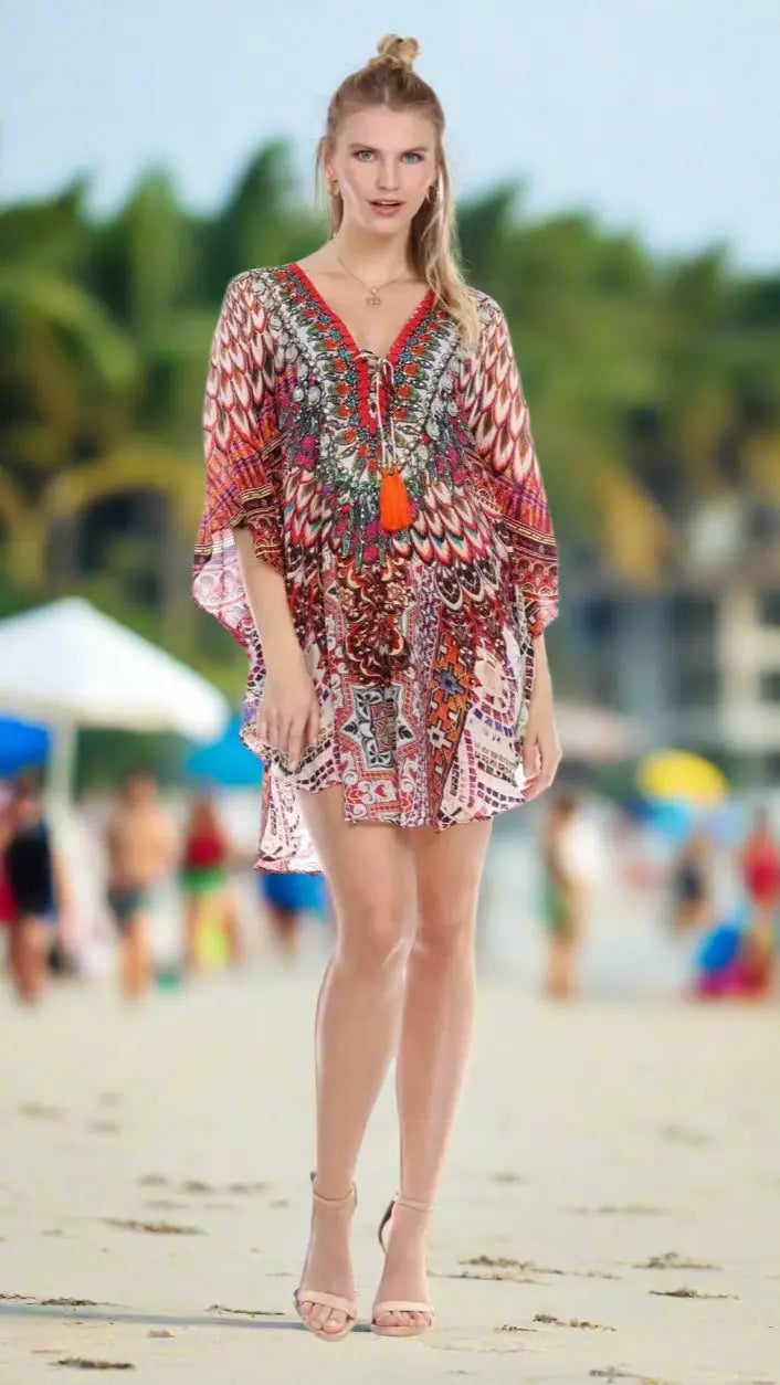 A woman stands on a beach in the Lamodaclothing Trendy and Comfy Short Kaftan Top, styled with nude heeled sandals and a necklace. Her hair is half-up in a top knot. The blurred background shows people, umbrellas, and palm trees.