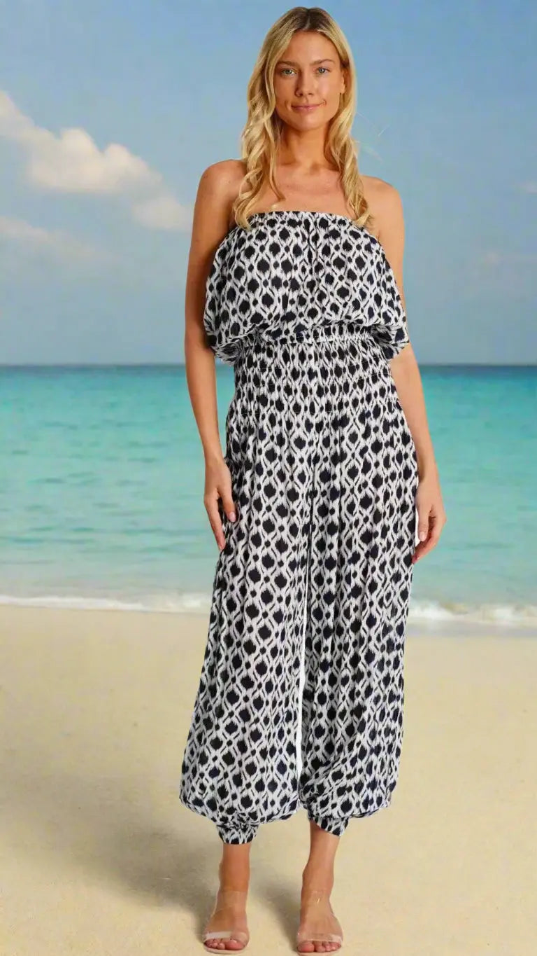 A woman with long blonde hair stands on a sandy beach, smiling in the sun. She wears La Moda Clothing's Tropical Jumpsuit—a strapless, black-and-white patterned piece with wide legs—paired with clear sandals by turquoise water and a blue sky.