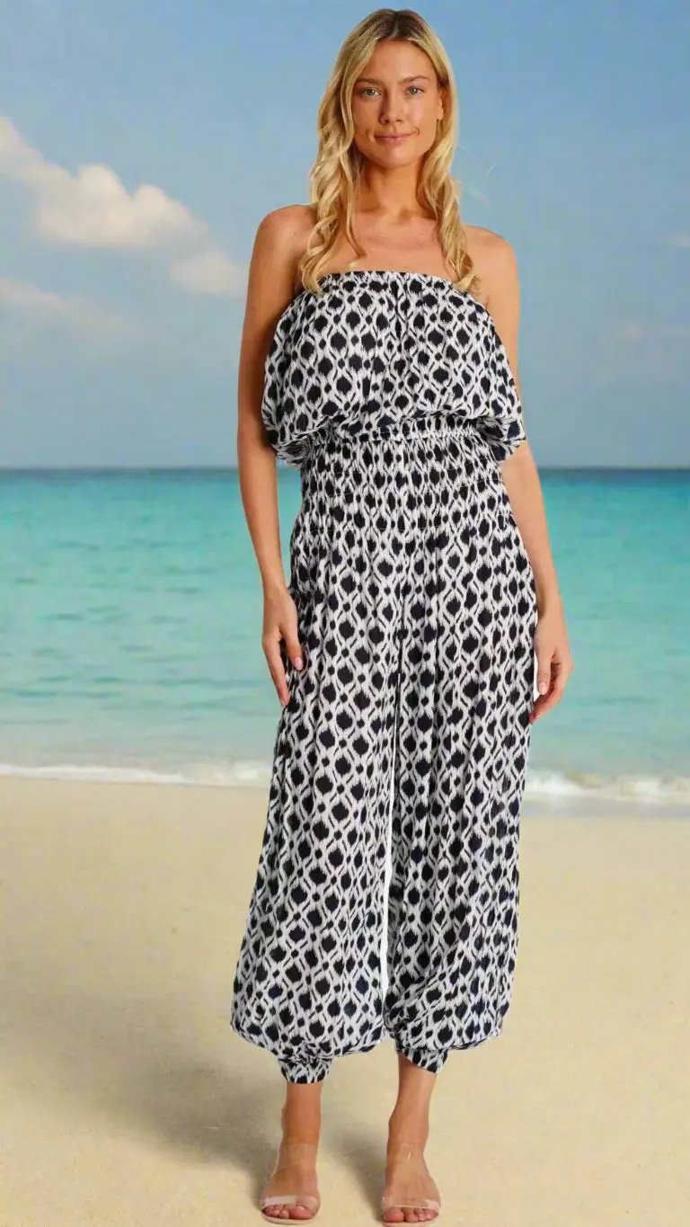 A woman with long blonde hair stands on a sandy beach, smiling in the sun. She wears La Moda Clothing's Tropical Jumpsuit—a strapless, black-and-white patterned piece with wide legs—paired with clear sandals by turquoise water and a blue sky.