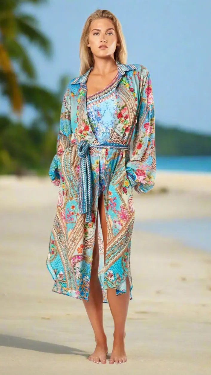 A woman stands barefoot on a sandy beach in the La Moda Clothings True Colors Easy Shirtdress Cover-Up, featuring colorful floral and geometric patterns over a blue swimsuit. Green palms and the sea blur in the background.
