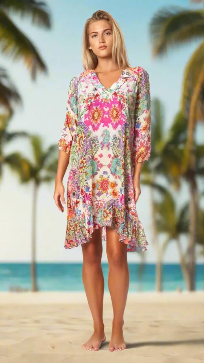A woman with blonde hair stands barefoot on a sandy beach, wearing the La Moda Clothings V Neck Mini Dress with Sleeves. Palm trees and a clear blue sky with ocean in the background create a tropical, relaxed vibe.