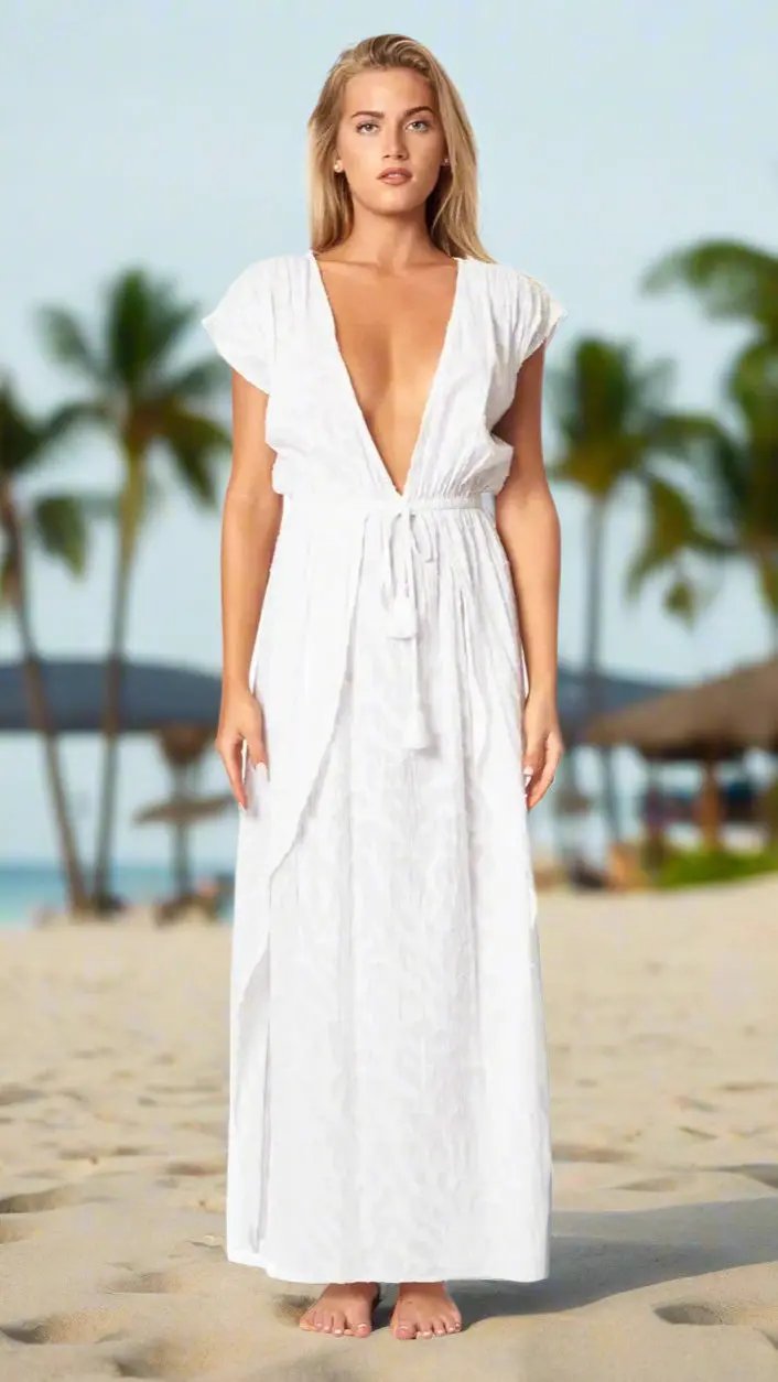 A woman stands barefoot on a sandy beach under palm trees, wearing the White Maxi Dress with Front Slits by La Moda Clothing. The elegant dress features short sleeves and a deep V-neckline, perfect for sunny days by the sea.