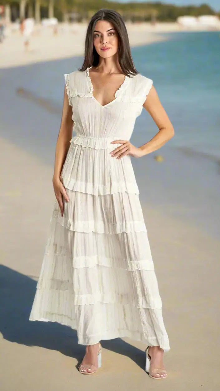 A woman with long brown hair poses by the shore in La Moda Clothing’s Tiered Maxi Dress in White for Coastal Stays, featuring a sleeveless V-neck, ruffle details, and strappy heels, with sandy beach and trees in the background.