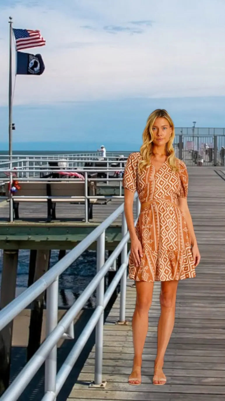 A woman in La Moda Clothings Womens Cutout Mini Dress stands on a wooden pier by the ocean, flanked by railings and benches, as U.S. and state flags wave nearby beneath a partly cloudy sky.