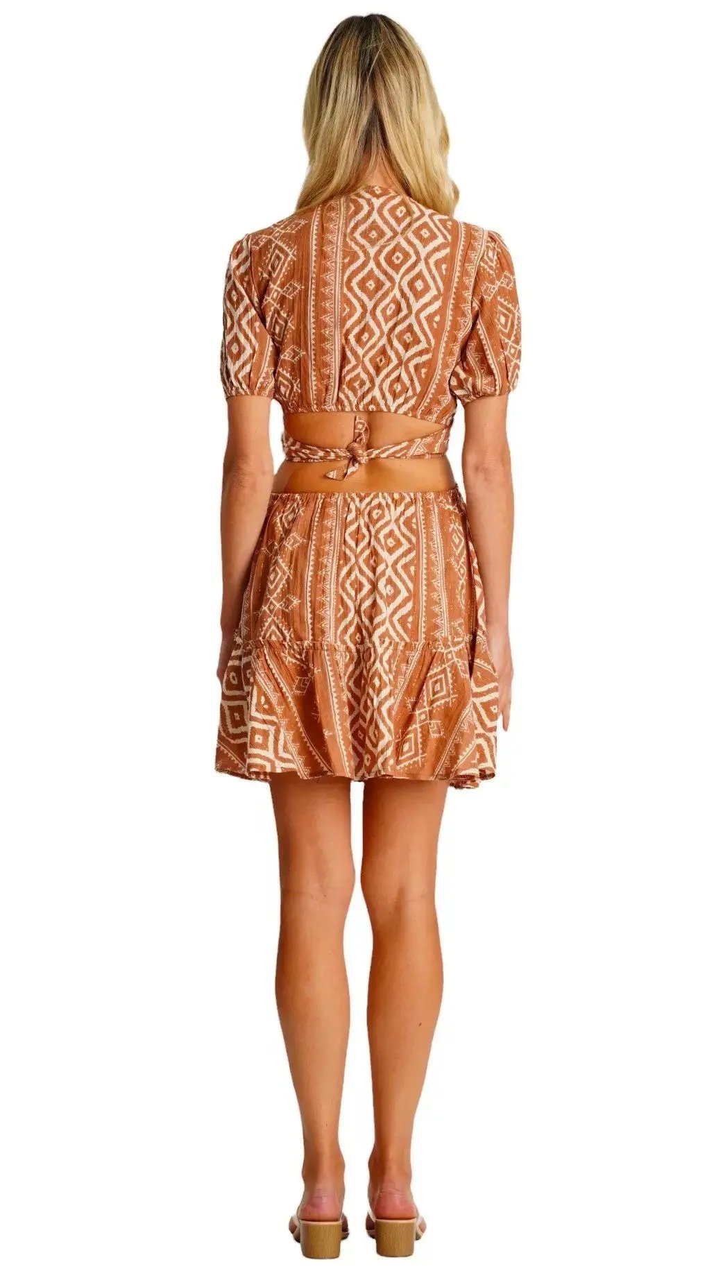 A woman with long blonde hair stands facing away in the La Moda Clothing Womens Cutout Mini Dress—a short-sleeve, brown and white patterned summer dress with a cutout back, tie detail, and deep V-neckline—paired with heeled sandals.