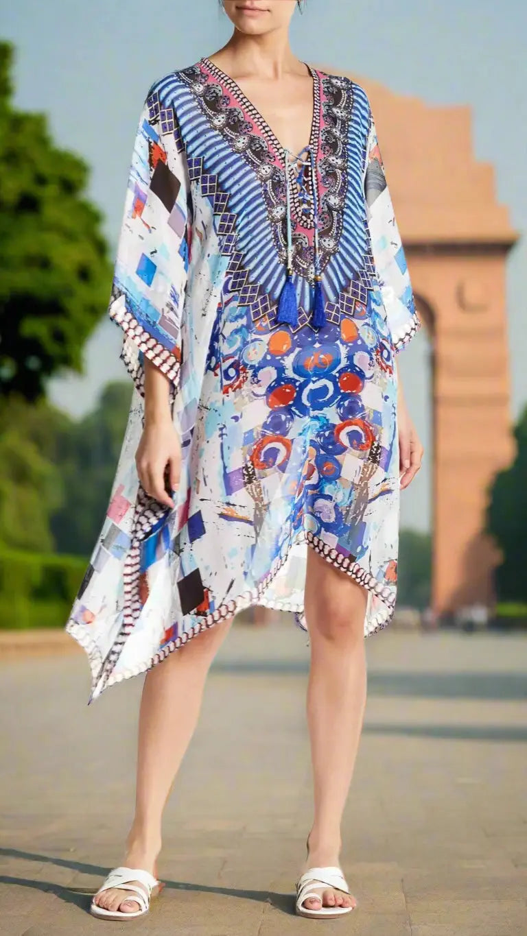 A woman outdoors near a monument wears the Women's Kaftan by La Moda Clothing, featuring blue, red, and white geometric and abstract patterns. She pairs it with white sandals and a necklace, her head slightly turned and face partially out of frame.