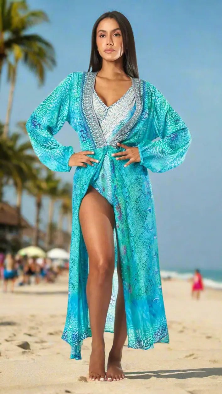A woman models La Moda Clothing’s Women's Maxi Wrap Dress for Sea Cruise Escape on a sunny beach, its blue and turquoise pattern and high slit flowing over her matching swimsuit, with palm trees blurred in the background.