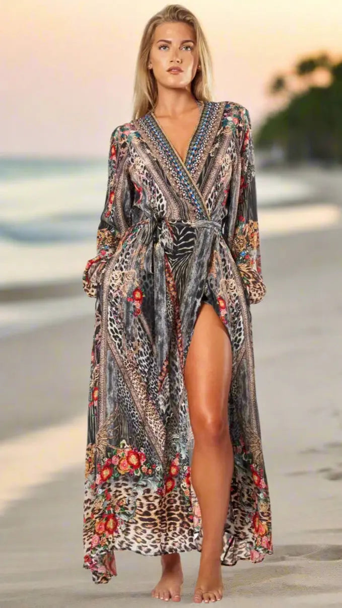 At sunset, a woman strolls barefoot on the beach in La Moda Clothing's Women's Maxi Wrap Dress for Summer Vacay—an animal-print, long-sleeve dress with floral patterns, deep V-neck, and high slit. Palm trees and ocean blur in the background.