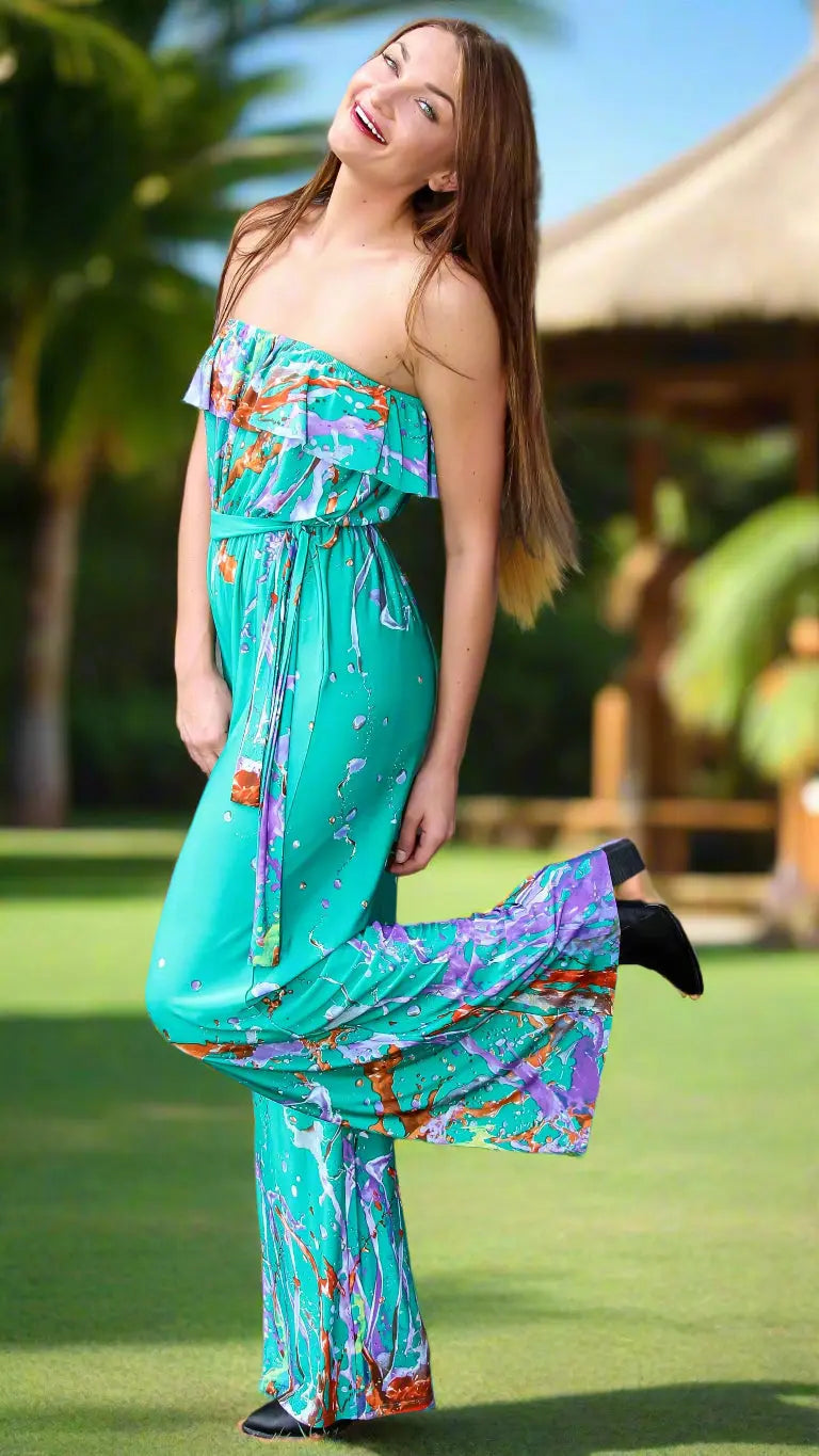 A smiling woman with long brown hair poses outdoors in the Lamodaclothing summer jumpsuit for women, featuring a sleeveless turquoise design with colorful patterns. She stands on grass, playfully lifting one leg, with palm trees and a thatched roof behind her.