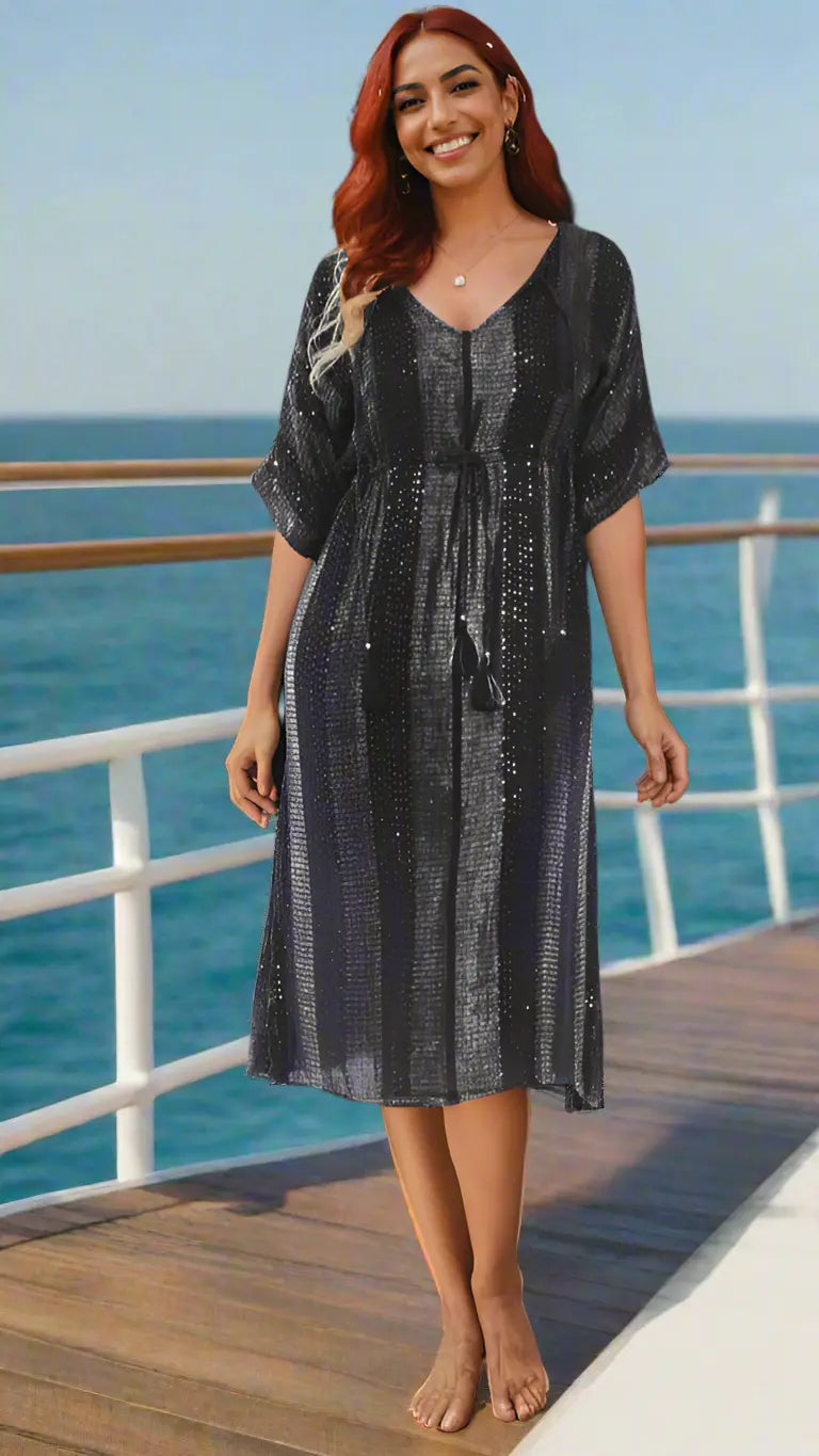 A smiling woman stands barefoot on a ship deck by the sea, wearing the Lamodaclothing Women's Swimsuit Cover up Beach Kaftan—a loose-fitting, knee-length black dress with sparkly stripes and a waist tie. Bright sky and blue ocean form the backdrop.