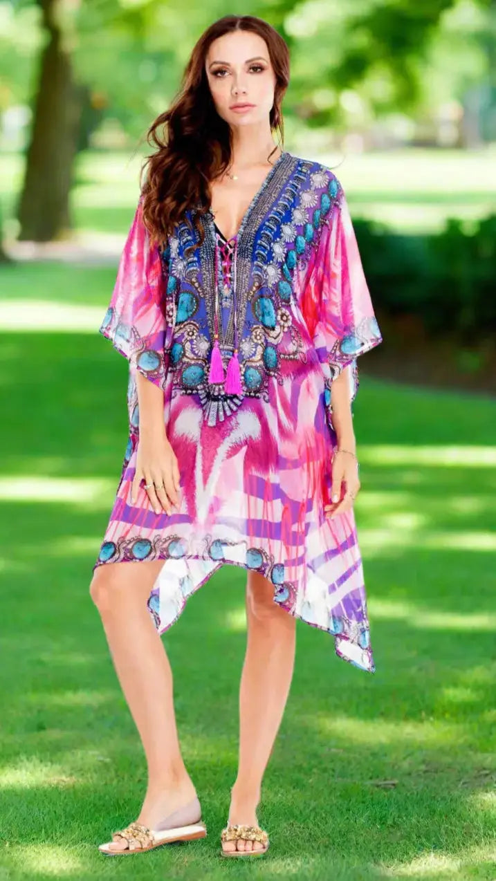 A woman with long wavy brown hair stands on park grass, wearing La Moda Clothing's Womens Short Luxury Caftan in vibrant pink and purple, paired with a tassel necklace and gold flat sandals. Green trees and sunlight blur in the background.