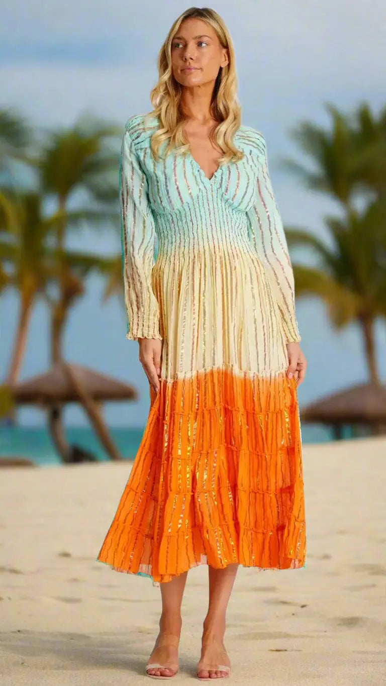 A woman with long blonde hair stands barefoot on a sandy beach, wearing the La Moda Clothing Womens Vintage Boho Maxi Dress, featuring aqua blue bodice, cream midsection, and bright orange skirt. Palm trees and beach umbrellas are in the background.