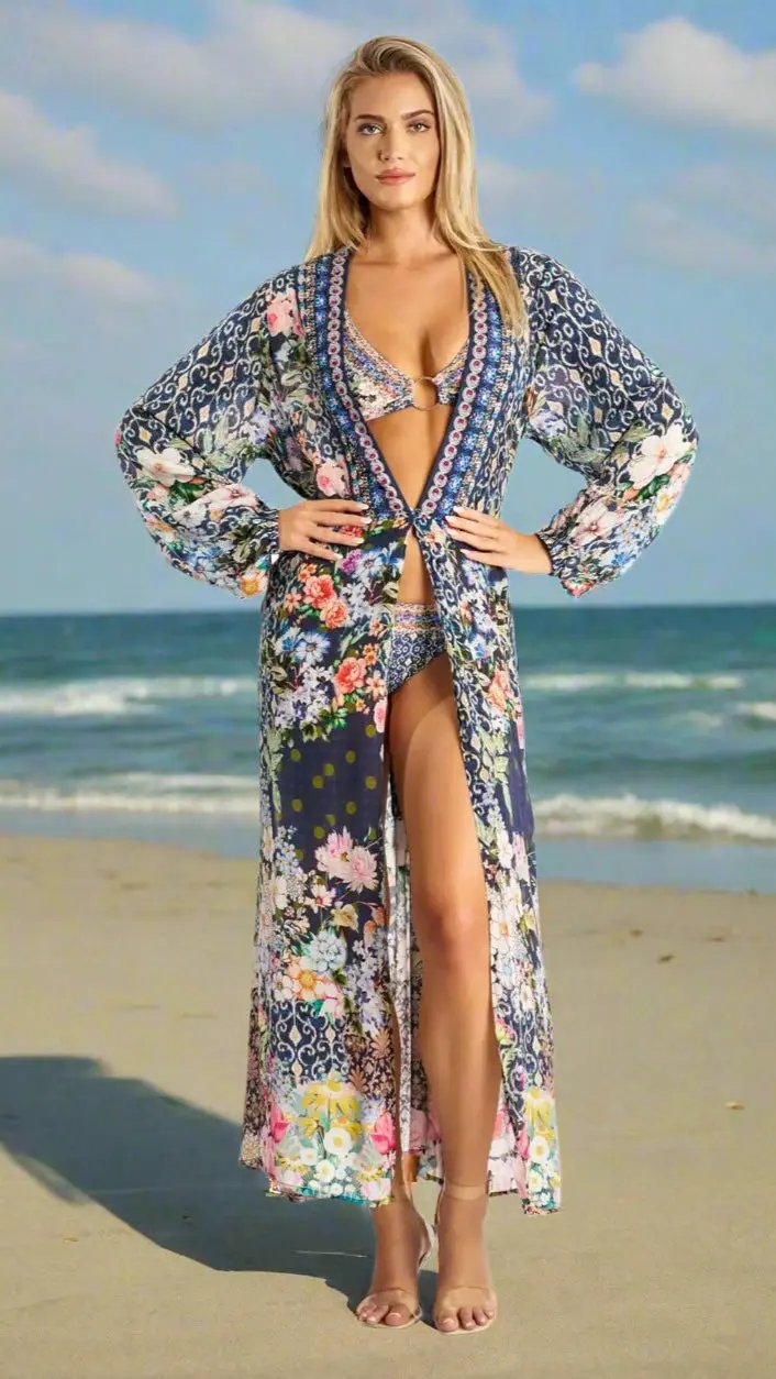 A woman stands confidently on a sandy beach in a La Moda Clothing wrap dress, showcasing effortless elegance and comfort beneath a blue sky with gentle waves in the background.