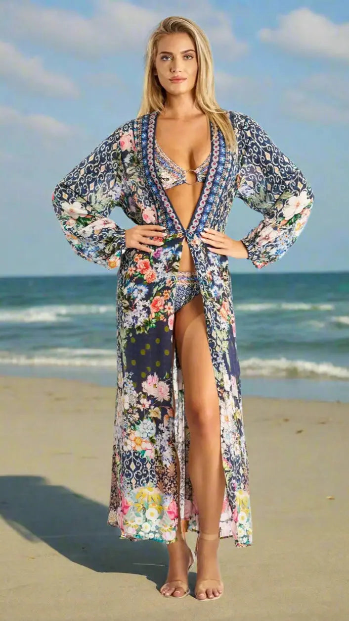 A woman stands confidently on a sandy beach in a La Moda Clothing wrap dress, showcasing effortless elegance and comfort beneath a blue sky with gentle waves in the background.