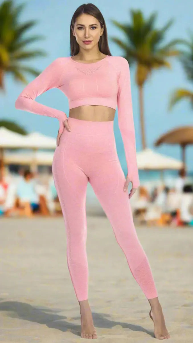 A woman stands barefoot on a sandy beach wearing the La Moda Clothings Women's Knitted Seamless Long Sleeve Top in light pink with matching high-waisted leggings. Palm trees and umbrellas with seated people appear blurred in the background.
