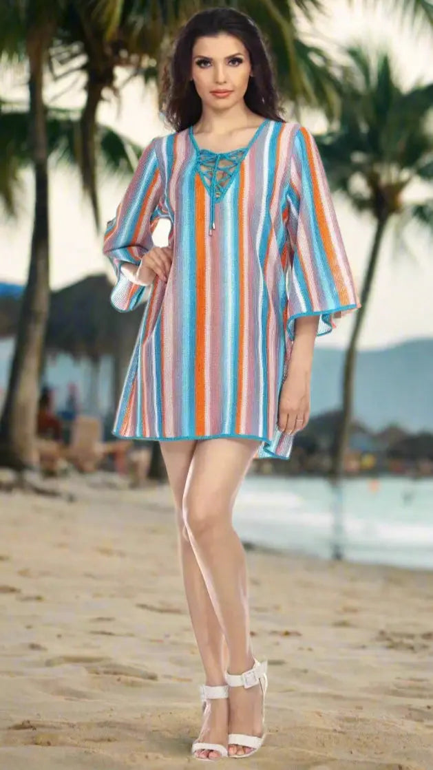 A woman stands on a sandy tropical beach with palm trees, wearing the Lamodaclothing Multi-Color Stripe Pattern Beach Cover-Up Tunic and white heeled sandals. The ocean shimmers serenely in the background.