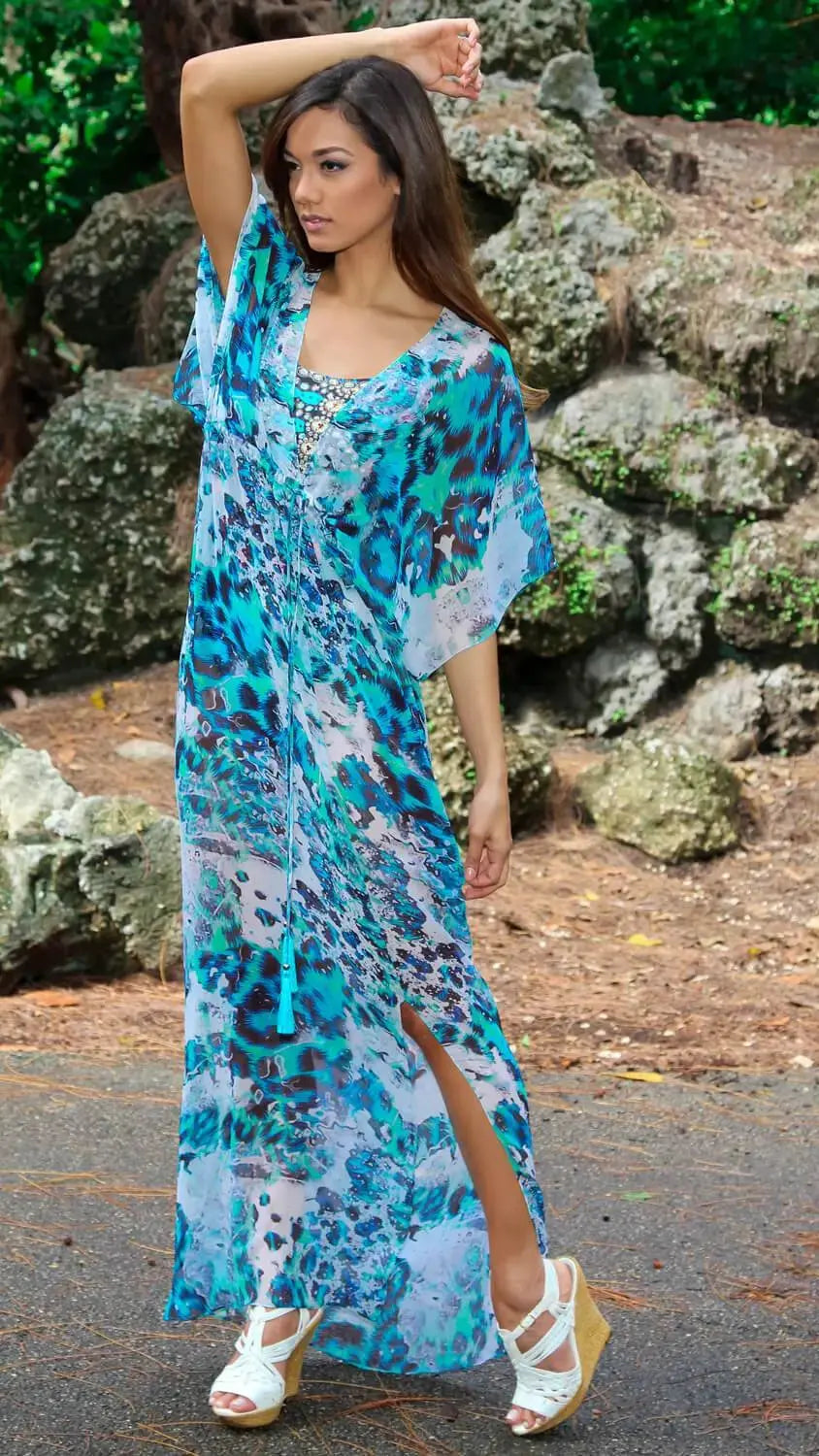 A woman poses outdoors among rocks and greenery, wearing the lamodaclothings Royal Jungle Printed Long Kaftan-Style Beachwear Cover Up in blue and turquoise animal print, paired with white wedge sandals.