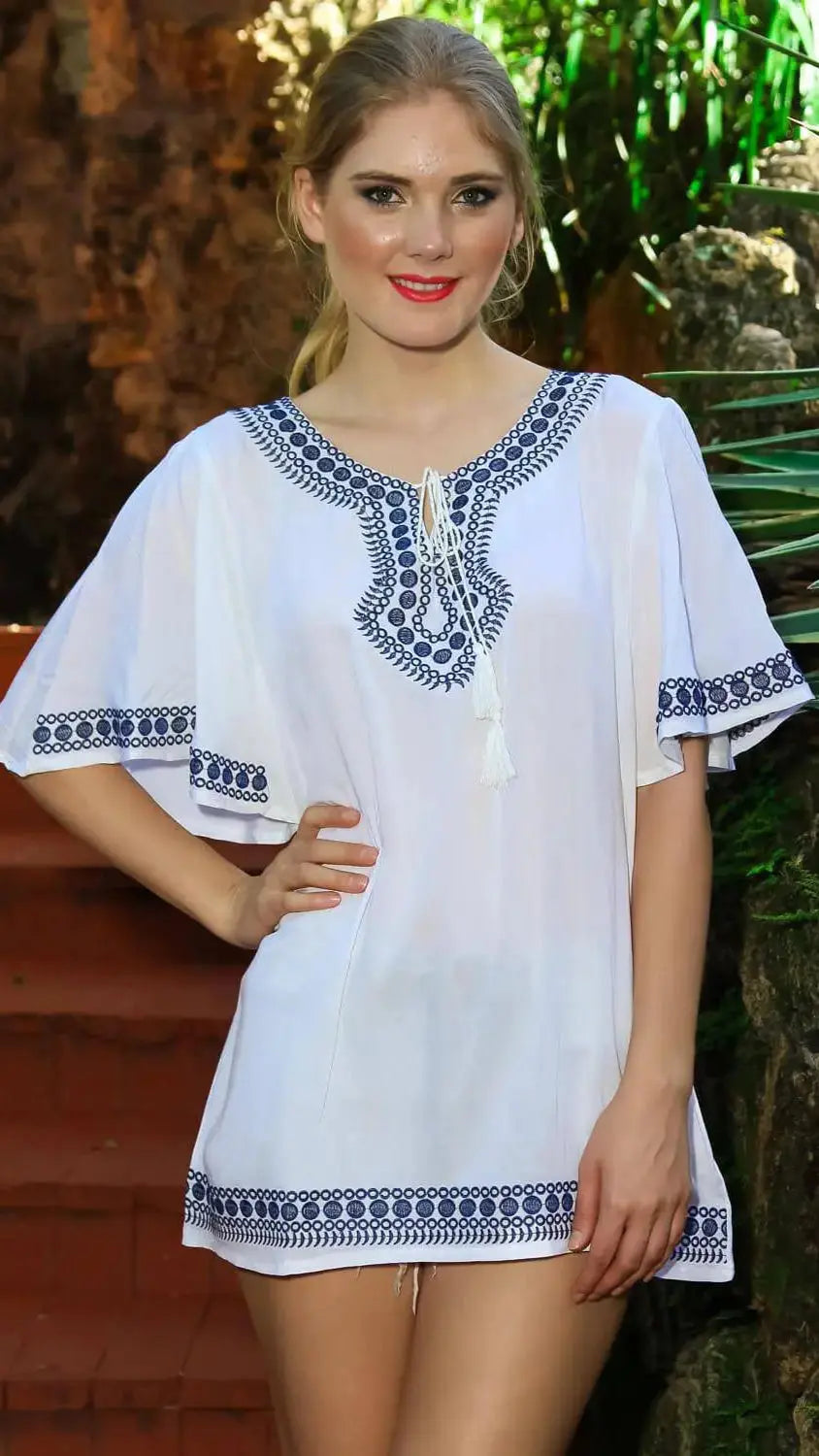 A smiling woman with blonde hair stands outdoors by rocky steps, wearing lamodaclothings' Embroidered Mini Tunic Cover Up for Beachwear—white with blue embroidery at the neckline, sleeves, and hem.