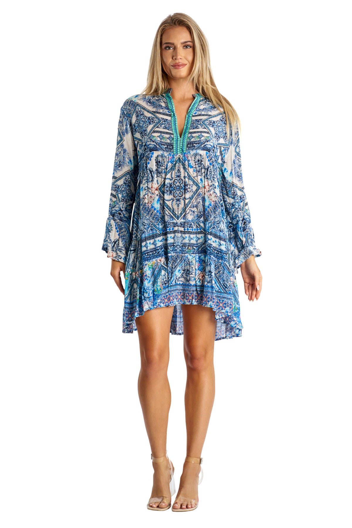 A woman with long blonde hair faces forward, wearing a blue patterned Spring Dress by La Moda Clothing. The loose-fitting dress has a green neckline detail, long sleeves, and is made from lightweight fabric. She pairs it with beige ankle-strap heels.