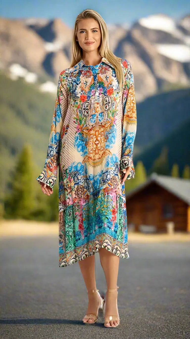 A woman with long blonde hair smiles outdoors in the La Moda Clothing A-Line Kaftan Collar Resort Dress, featuring a colorful, loose fit and long sleeves against a scenic background of mountains, trees, and a wooden cabin under a blue sky.