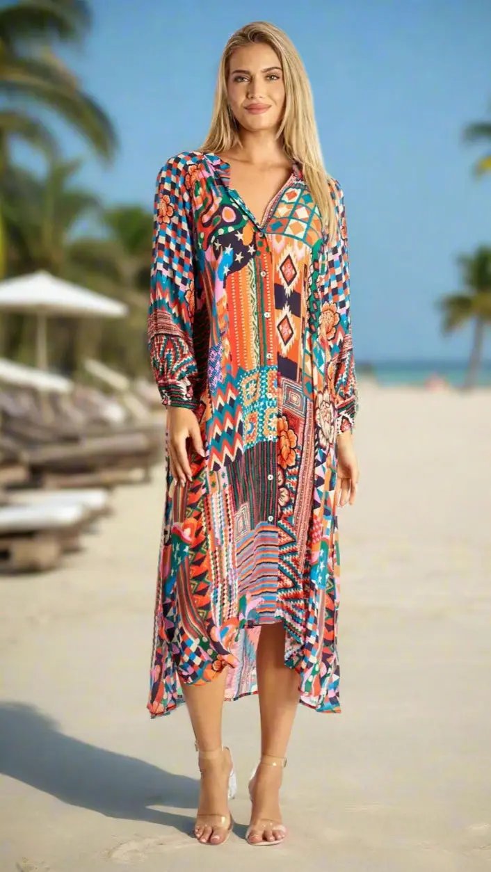 A smiling blonde woman stands barefoot on a sandy beach in a La Moda Clothing A-Line Kaftan Collar Shirt Dress, featuring long sleeves and vibrant geometric and floral patterns. Palm trees and empty lounge chairs are behind her under a clear blue sky.
