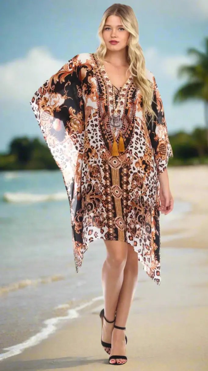 A woman with long blonde hair models the Caftans in Animal Print by La Moda Clothings—a loose dress with leopard and ornate patterns in brown, gold, and black—as she strolls barefoot along a sandy beach lined with palm trees.
