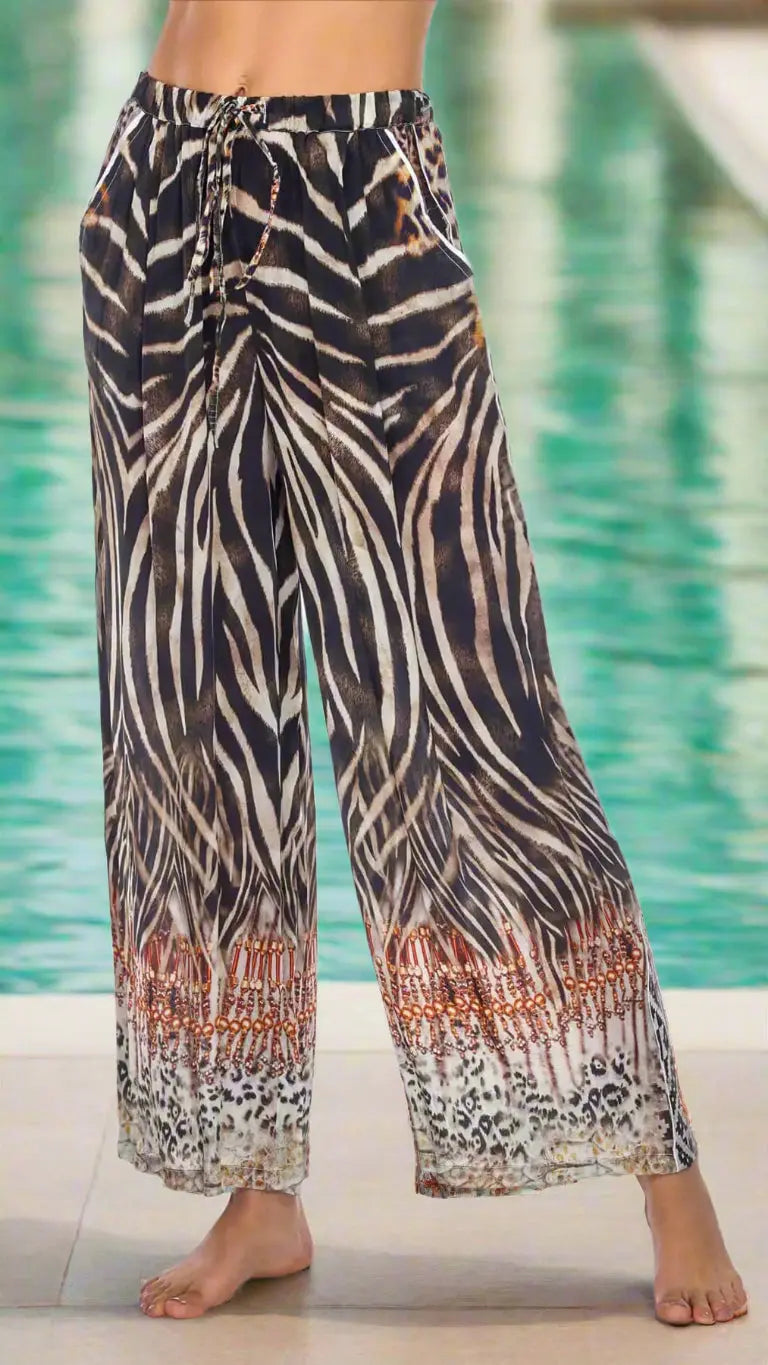 A person stands barefoot by a pool in La Moda Clothing's Animal Print Pants, featuring a high-waisted fit, black and white zebra pattern, leopard spots at the cuffs, and rusty orange accents. The blurred background highlights the bold design.