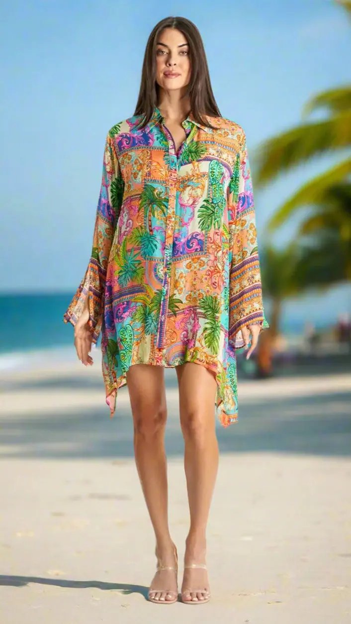 A woman with straight brown hair and neutral makeup stands on a sandy beach with palm trees, wearing the La Moda Clothings Beach Resort Cruise Easy Shirtdress Cover-Up and transparent heeled sandals.