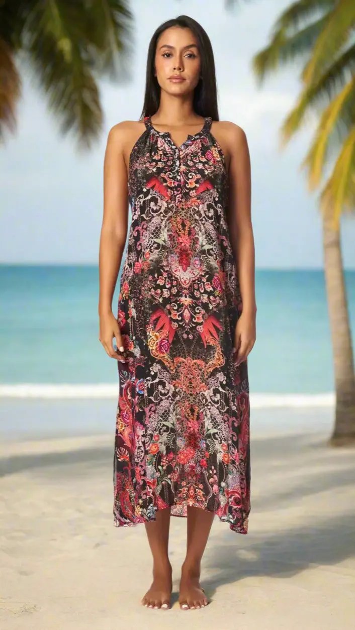 A woman with long dark hair stands barefoot on a sandy beach, wearing La Moda Clothing’s Beach Resort & Cruise Vacation Dress—an ankle-length, sleeveless dress with a vibrant red, pink, and black floral pattern—gazing calmly at the camera.
