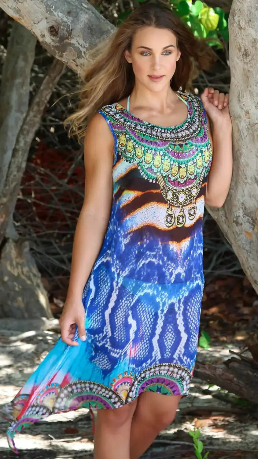 A woman stands outdoors among tree trunks, wearing the lamodaclothings Blue Python Midi Dress Beach Cruise Cover-Up. Sunlight filters through the trees as she gazes gently at the camera, her long wavy brown hair cascading over her shoulders.