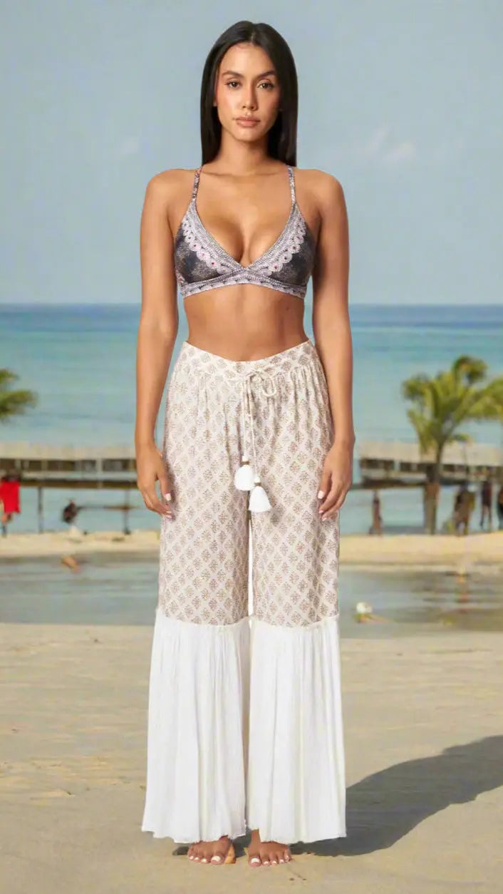 A woman stands barefoot on a sandy beach wearing the Boho Wide Leg Pant by La Moda Clothing—a high-waisted, wide-leg white pant with a patterned upper half and tassel tie. Palm trees, a pier, and calm blue water appear in the background under a clear sky.
