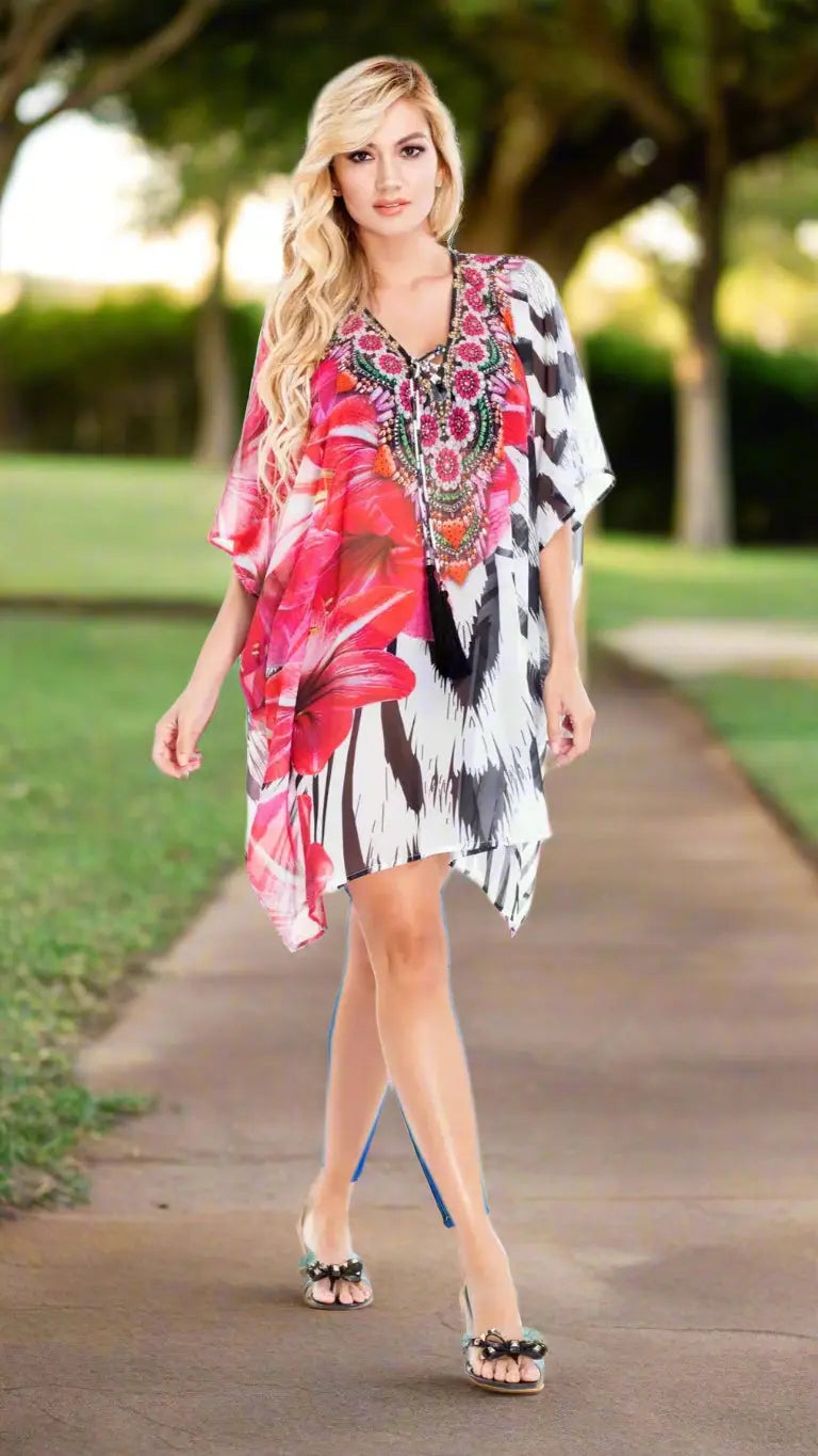 A woman with long blond hair wears the La Moda Clothing Caftan for Women, featuring a vibrant red floral and black-and-white pattern with a beaded neckline and tassel details. She pairs it with clear heeled sandals in a sunlit park.